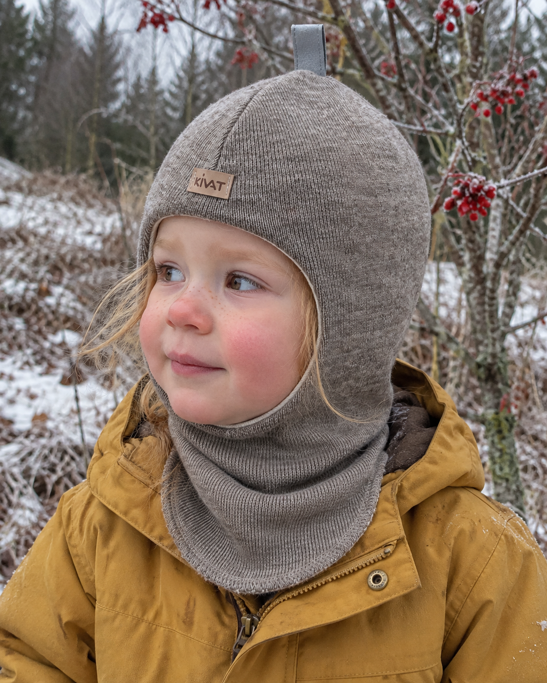 Liten jente i beige Kivat-balaclava i ull med liten Kivat-lapp, ute en frostmorgen
