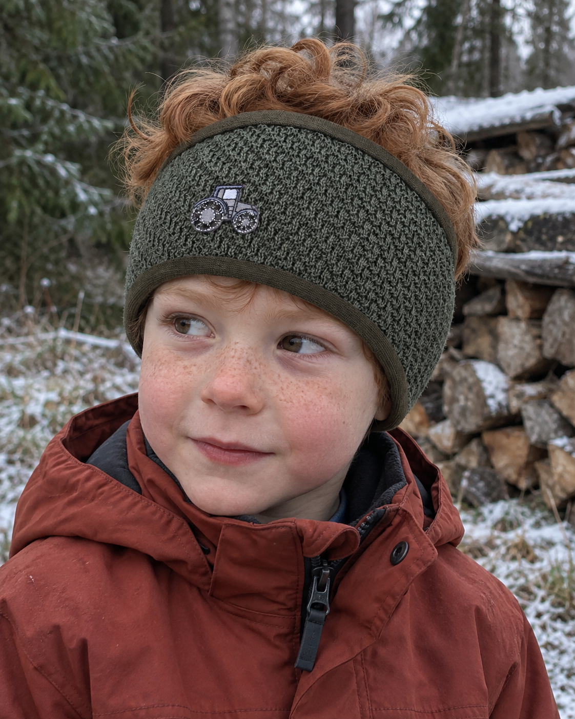 Gutt med grønt Kivat-pannebånd i ull med traktor-motiv, ute ved en vedstabel i frost