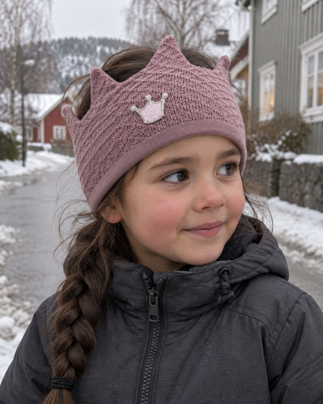 Jente med støvrosa Kivat-pannebånd i ull formet som en krone med kronemotiv, ute i en vintergate med snøslaps