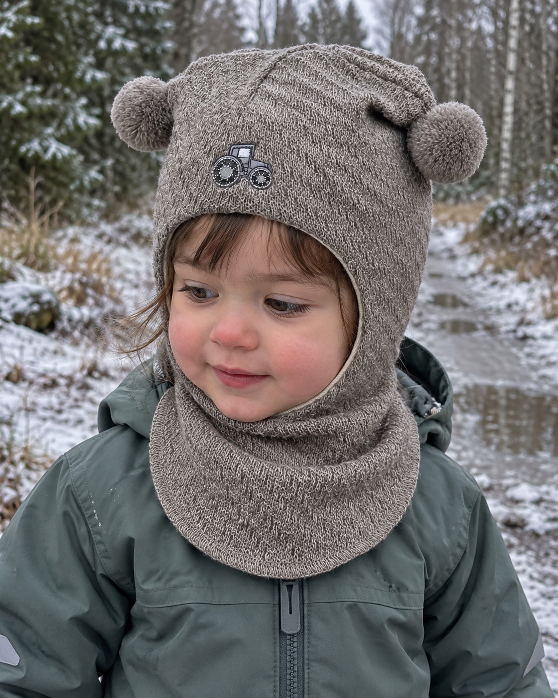 Liten jente i beige Kivat-balaclava i ull med traktor-motiv og pomponger, ute i vinterskog