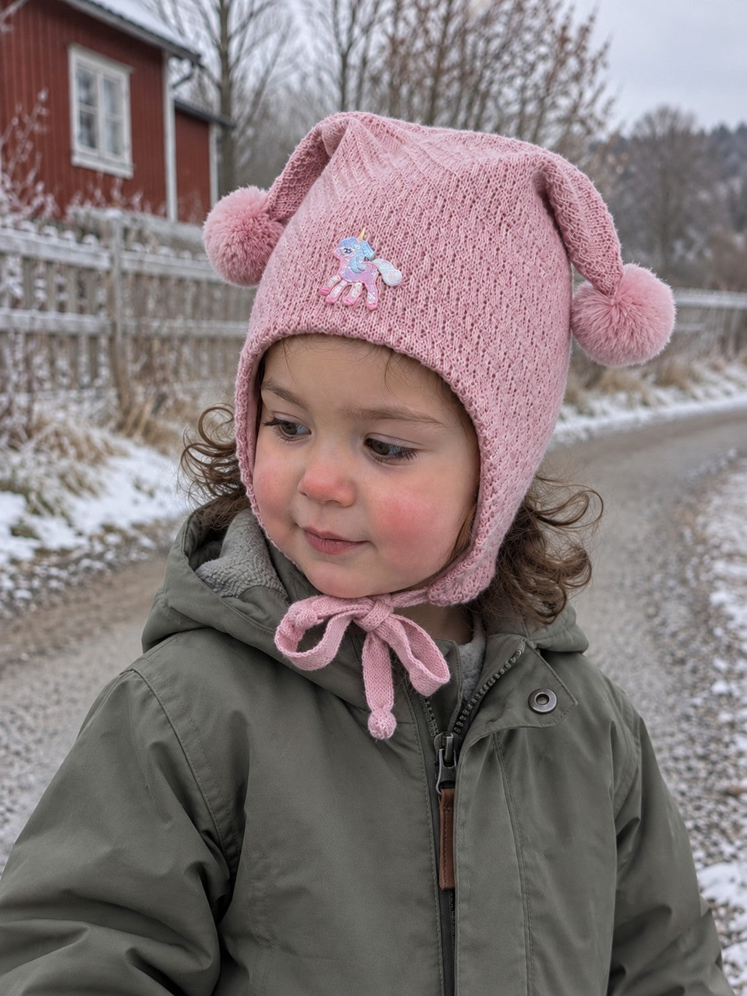 Lite barn i rosa Kivat-knytelue i ull med enhjørning-motiv og to pomponger, ute på en frostig bygdevei