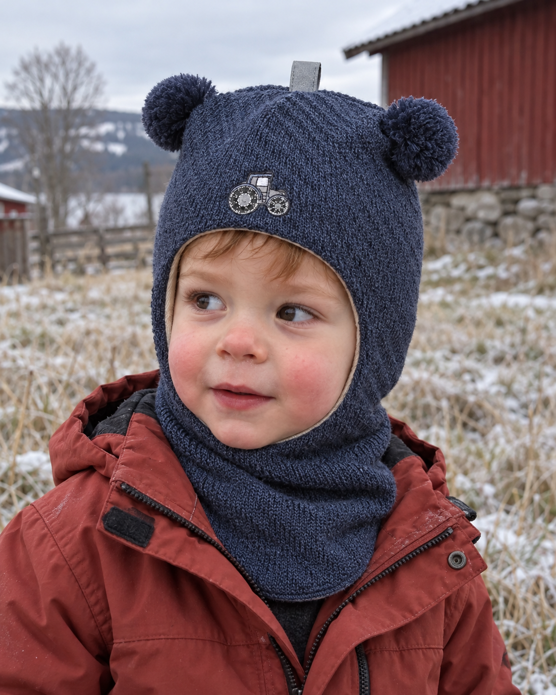 Liten gutt i blå melert Kivat-balaclava i ull med traktor-motiv og pomponger, ute en kald vinterdag