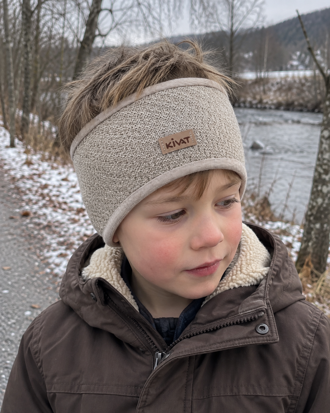 Gutt med Kivat-pannebånd i beige ull med Kivat-lapp, ute langs en elv i frost