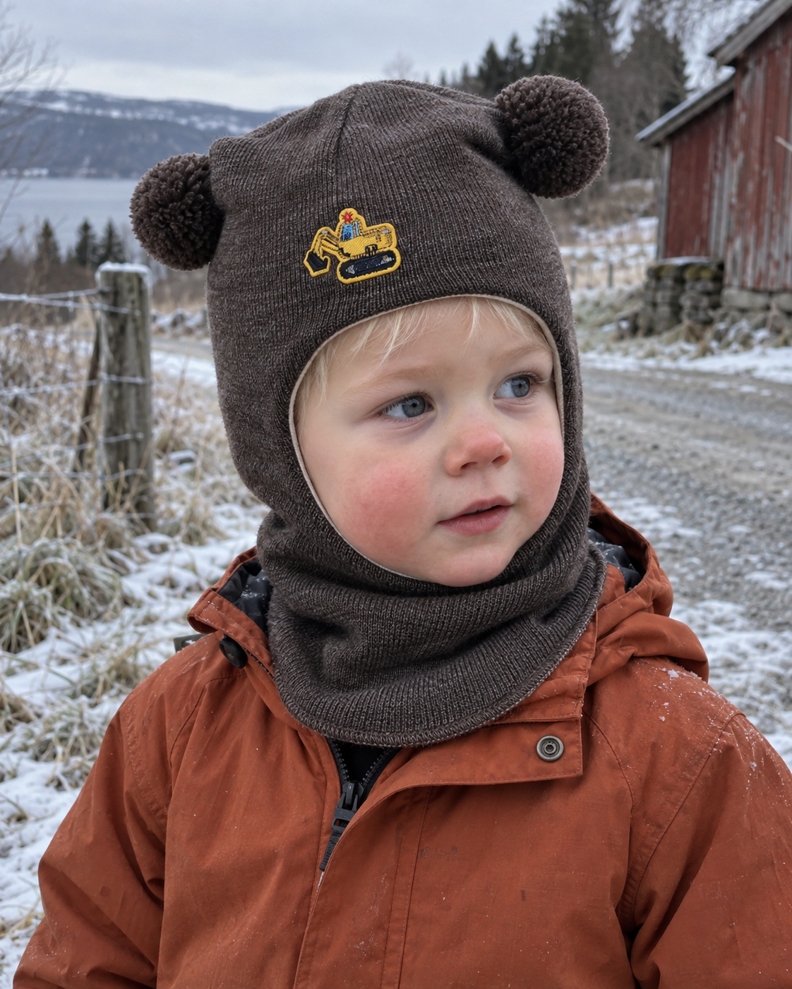 Liten gutt i brun melert Kivat-balaclava i ull med gravemaskin-motiv og pomponger, ute en kald vinterdag