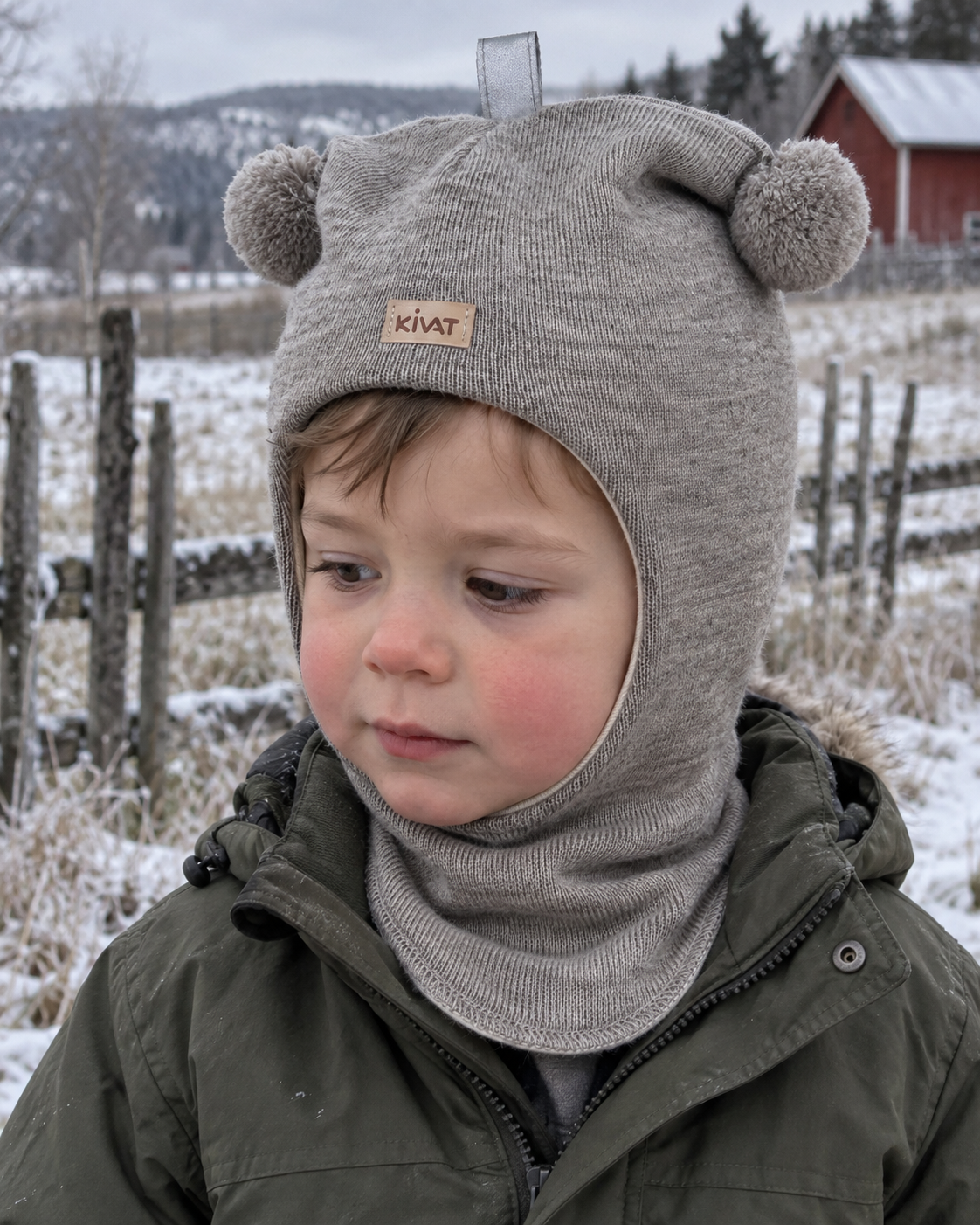 Liten gutt i beige Kivat-balaclava i ull med pomponger og Kivat-lapp, ute i rimfrost