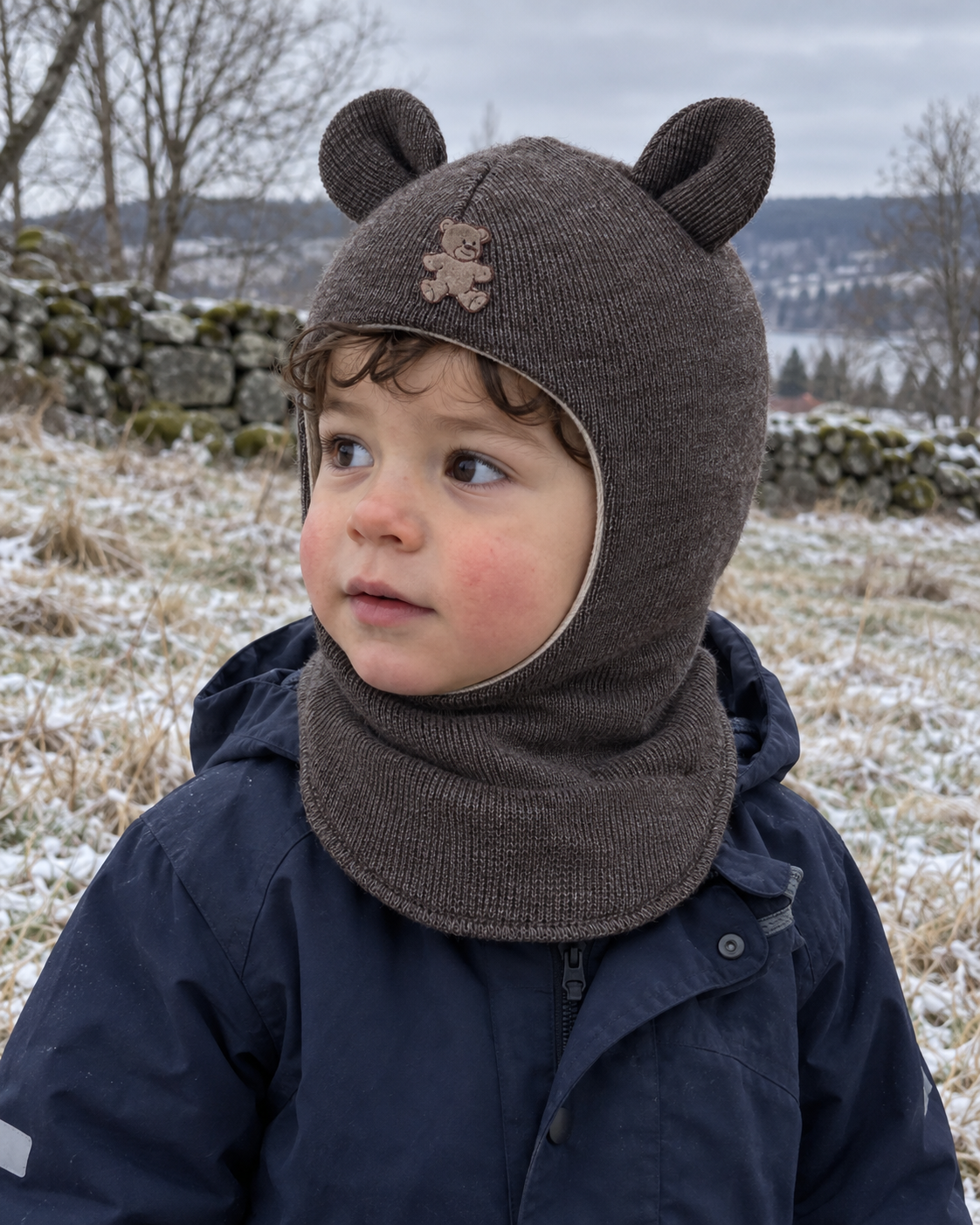Liten gutt i brun melert Kivat-balaclava i ull med bamseører, ute en kald vinterdag