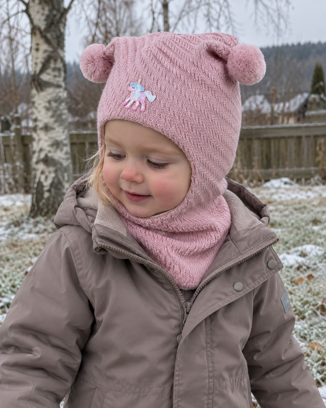 Liten jente i rosa Kivat-balaclava i ull med enhjørning og to pomponger, ute en kald vinterdag