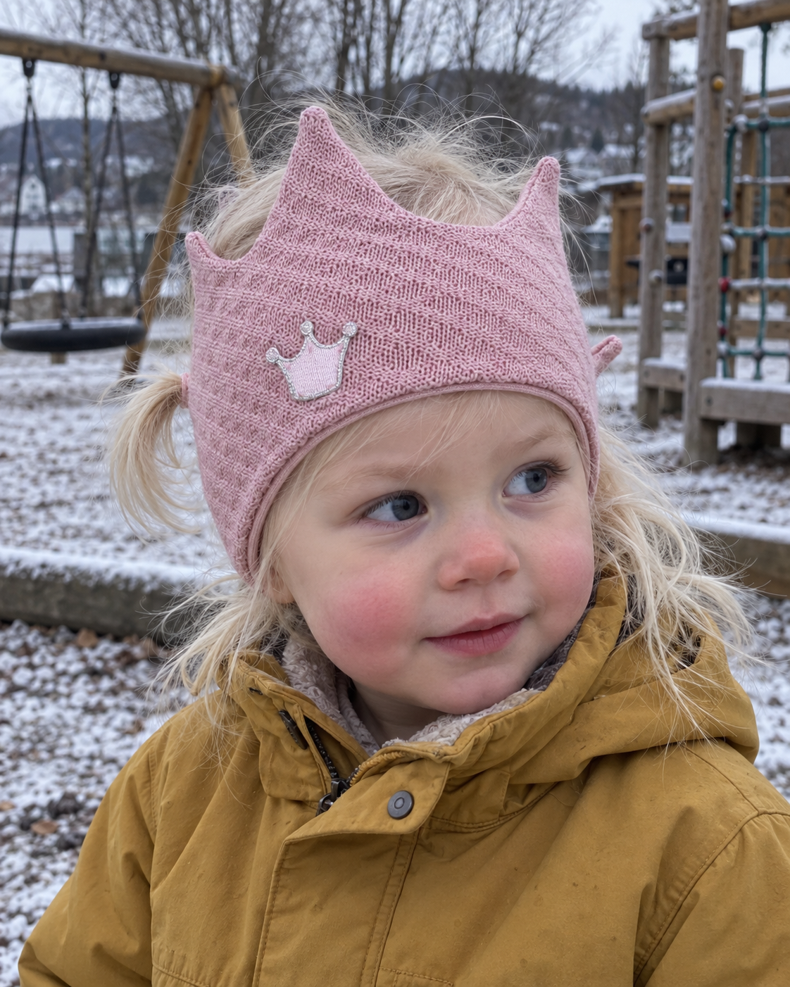 Liten jente med rosa Kivat-pannebånd i ull formet som en krone med kronemotiv, ute på en lekeplass med rim