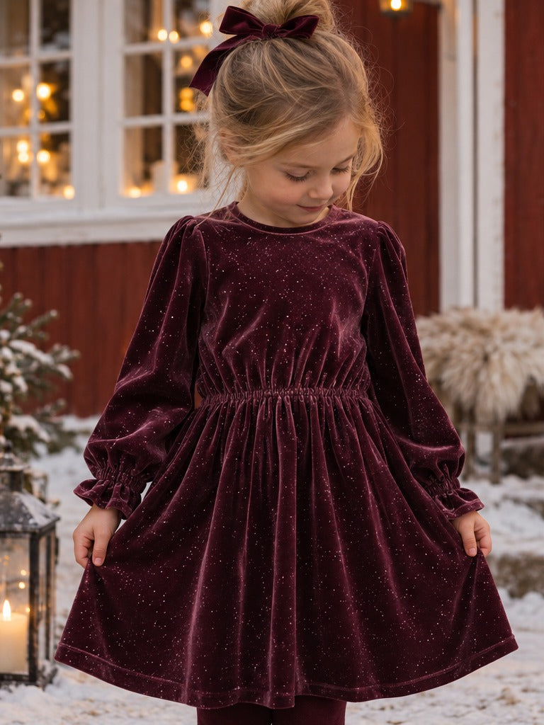 Jente i Name it Fricya velurkjole i burgunder ved julepyntet gårdshus i snøen