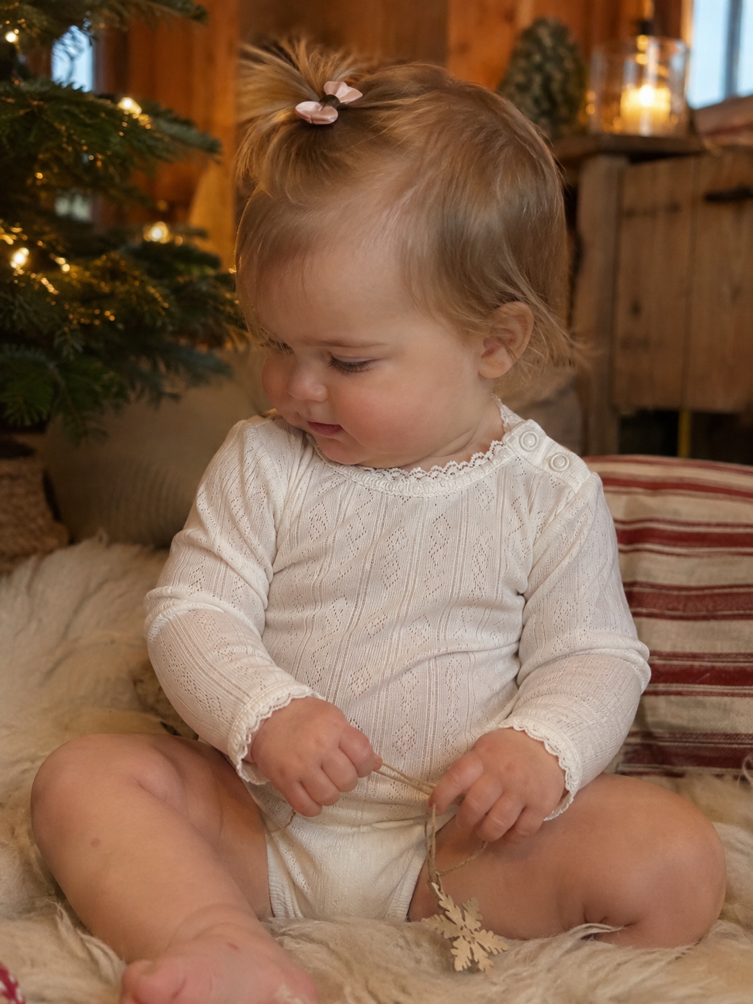Baby i Joha body i ull og silke med blonder sitter på saueskinn ved juletreet