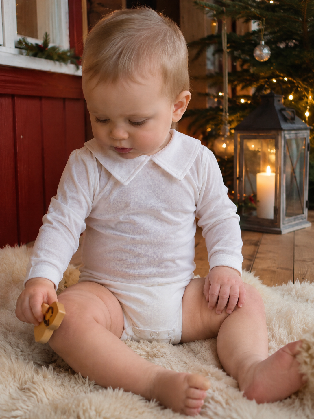 Babygutt i Joha hvit body med krage sitter på saueskinn i julepyntet gårdshus